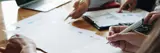 Three People Working At A Desk With Laptops Paperwork And Pens In Hand