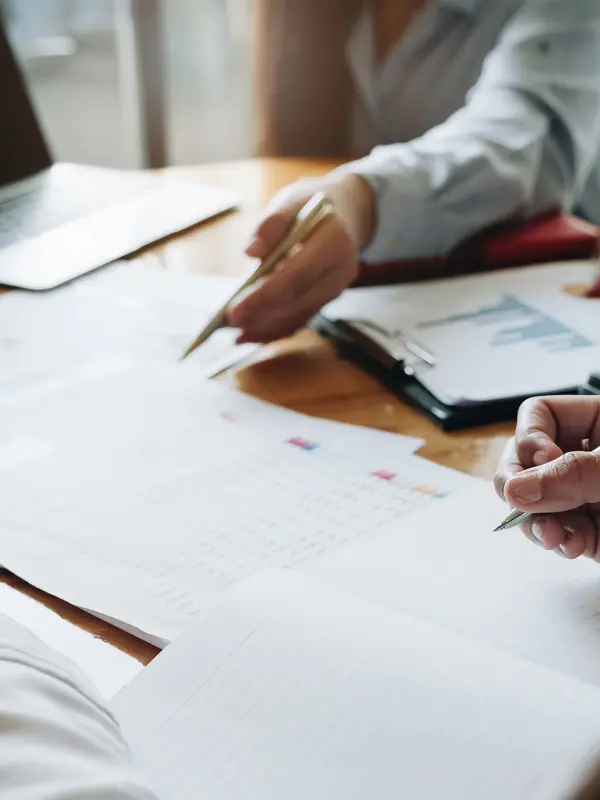Three People Working At A Desk With Laptops Paperwork And Pens In Hand